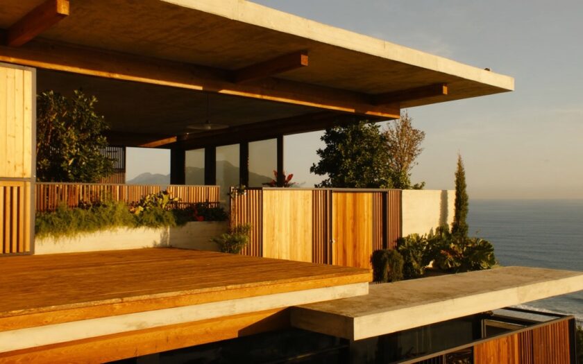Varanda profunda de uma casa, com deque e painéis de madeira, laje de concreto e plantas, voltada para o mar ao entardecer